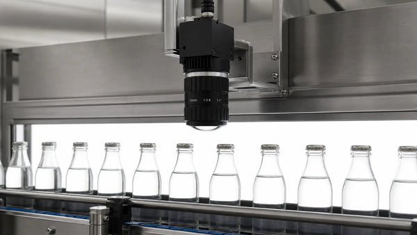 A machine vision camera with a C-mount lens inspecting backlit glass bottles on a conveyor